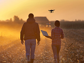 Frau mit einem Laptop in der Hand und Mann gehen während der Dämmerung in einem Stoppelfeld und eine Drohne fliegt im Hintergrund.